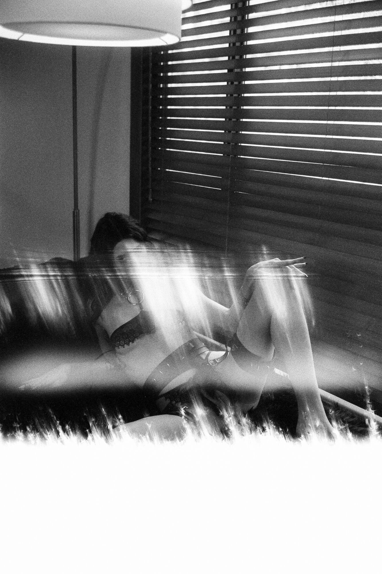 Black and white long-exposure portrait with motion blur, figure reclining against venetian blinds
