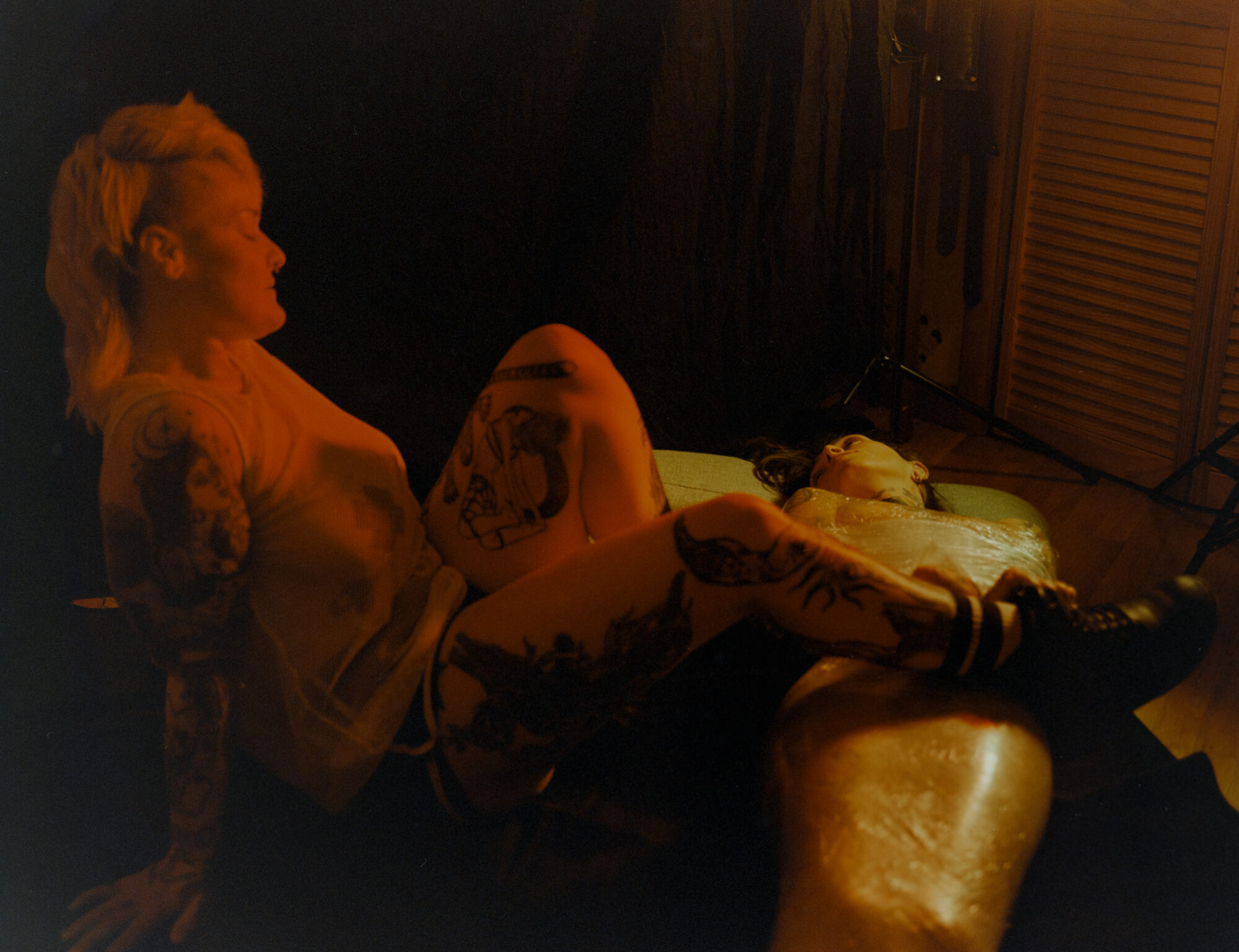 Warm amber-toned portrait of a tattooed figure, J, reclining in profile, deep shadows and rich golden light with their leg overtop of S, mummified in saran wrap. This was S's first experience being bound in plastic.
