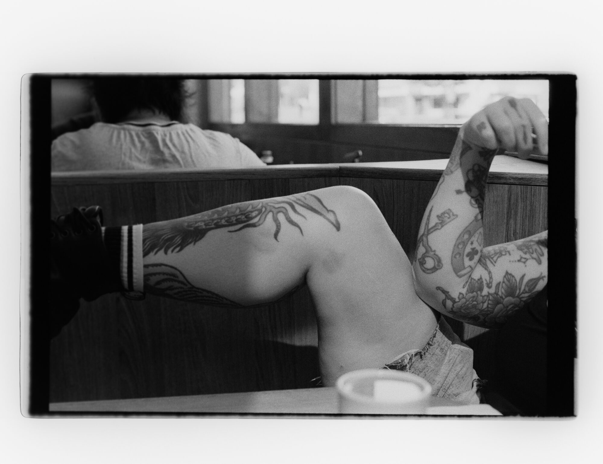Black and white diner scene, figure turned away showing tattooed arms with squid and floral designs