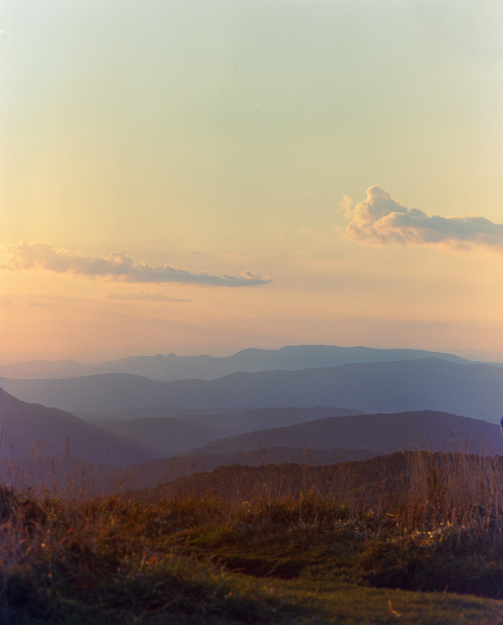Layered mountain ridges receding into haze at sunset, warm golden light on grass in the foreground, soft pink and blue sky taken from Max Patch, North Carolina.