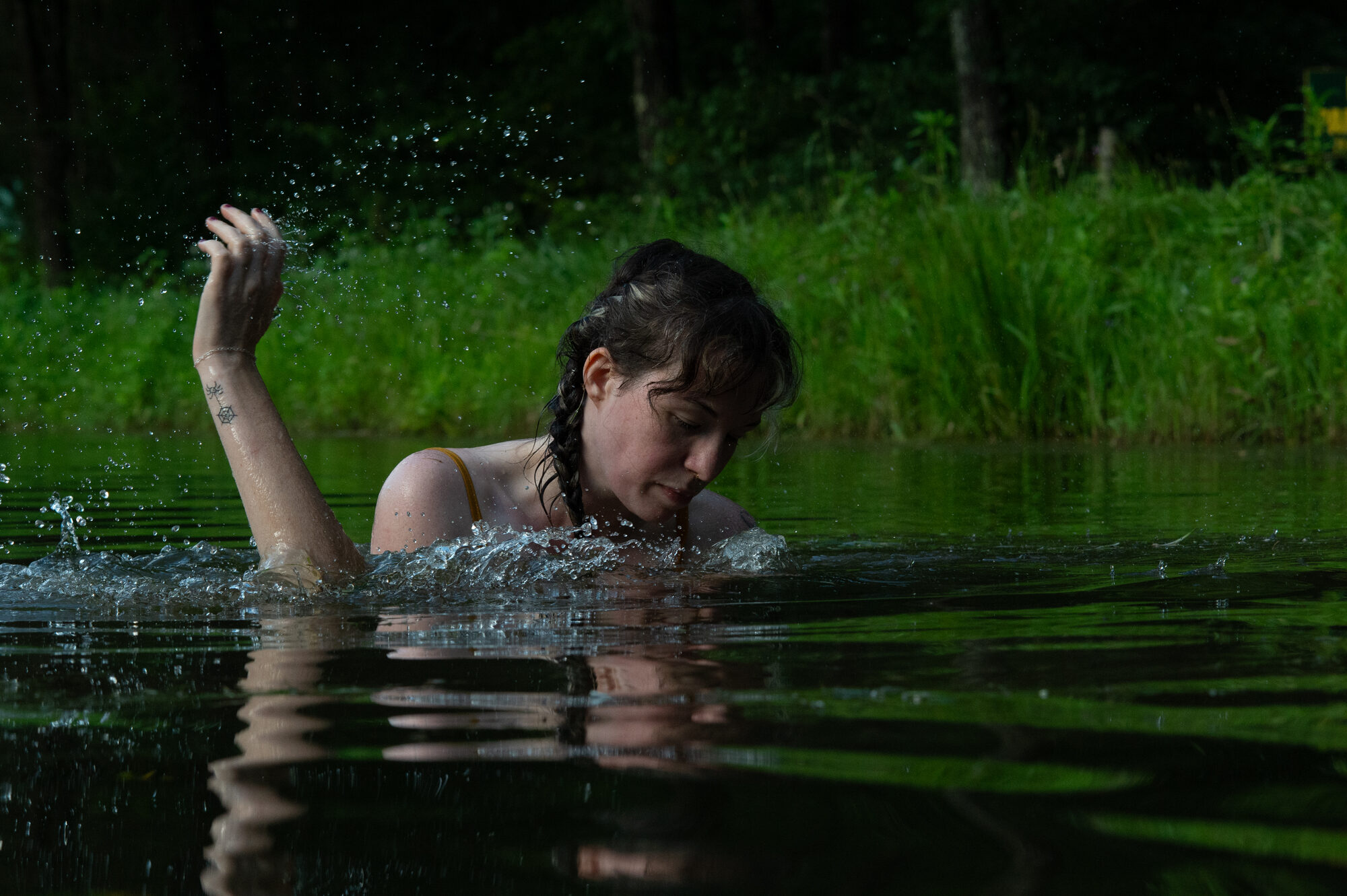 A figure splashing in a green lake with arm raised and water droplets, lush foliage behind. Part of a consensual drowning scene with the bottom's legs tied in an agura.