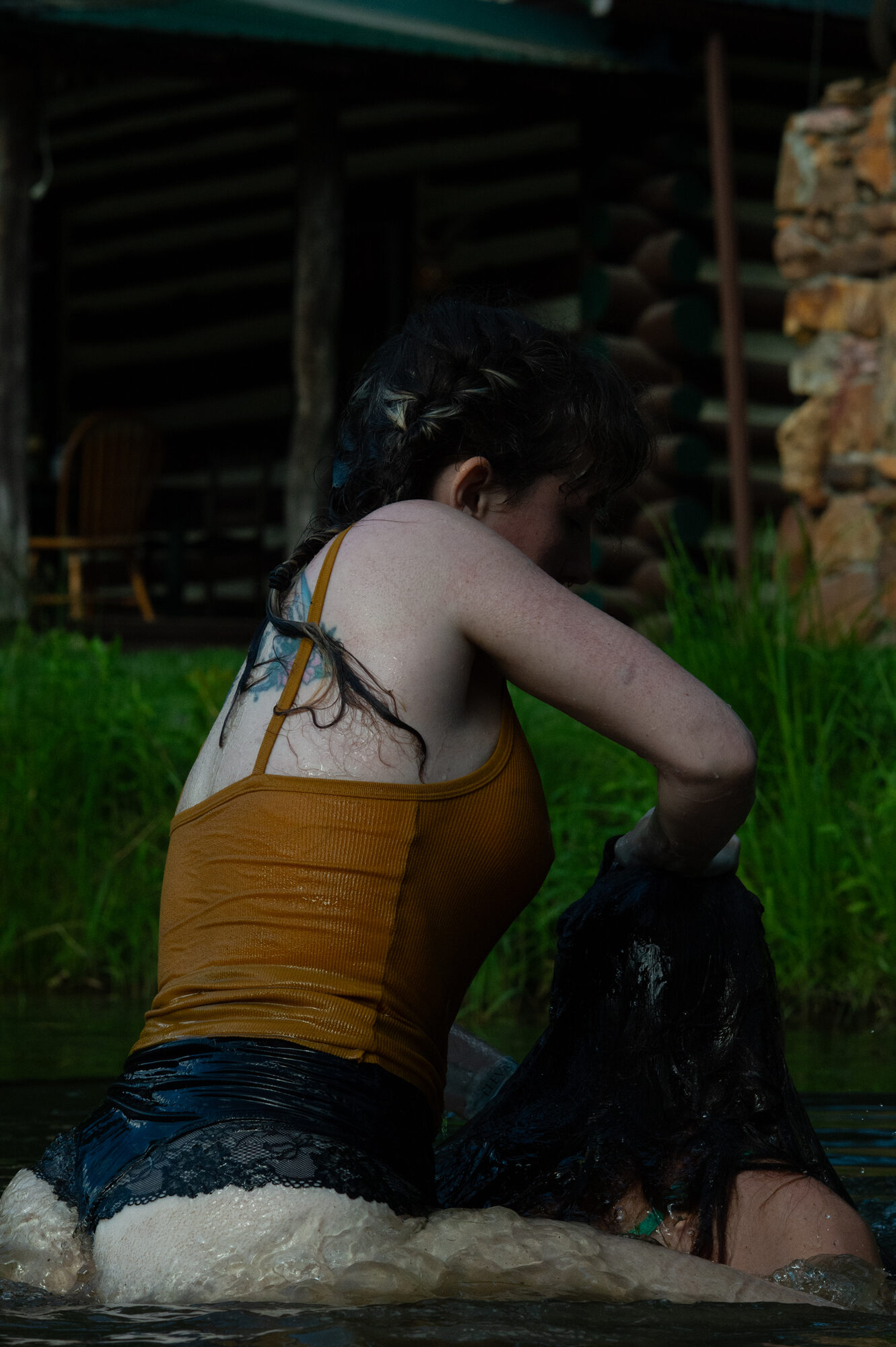Figure from behind wading in a lake wearing a yellow tank top, log cabin on the far shore. Part of a consensual drowning scene with the bottom's legs tied in an agura.