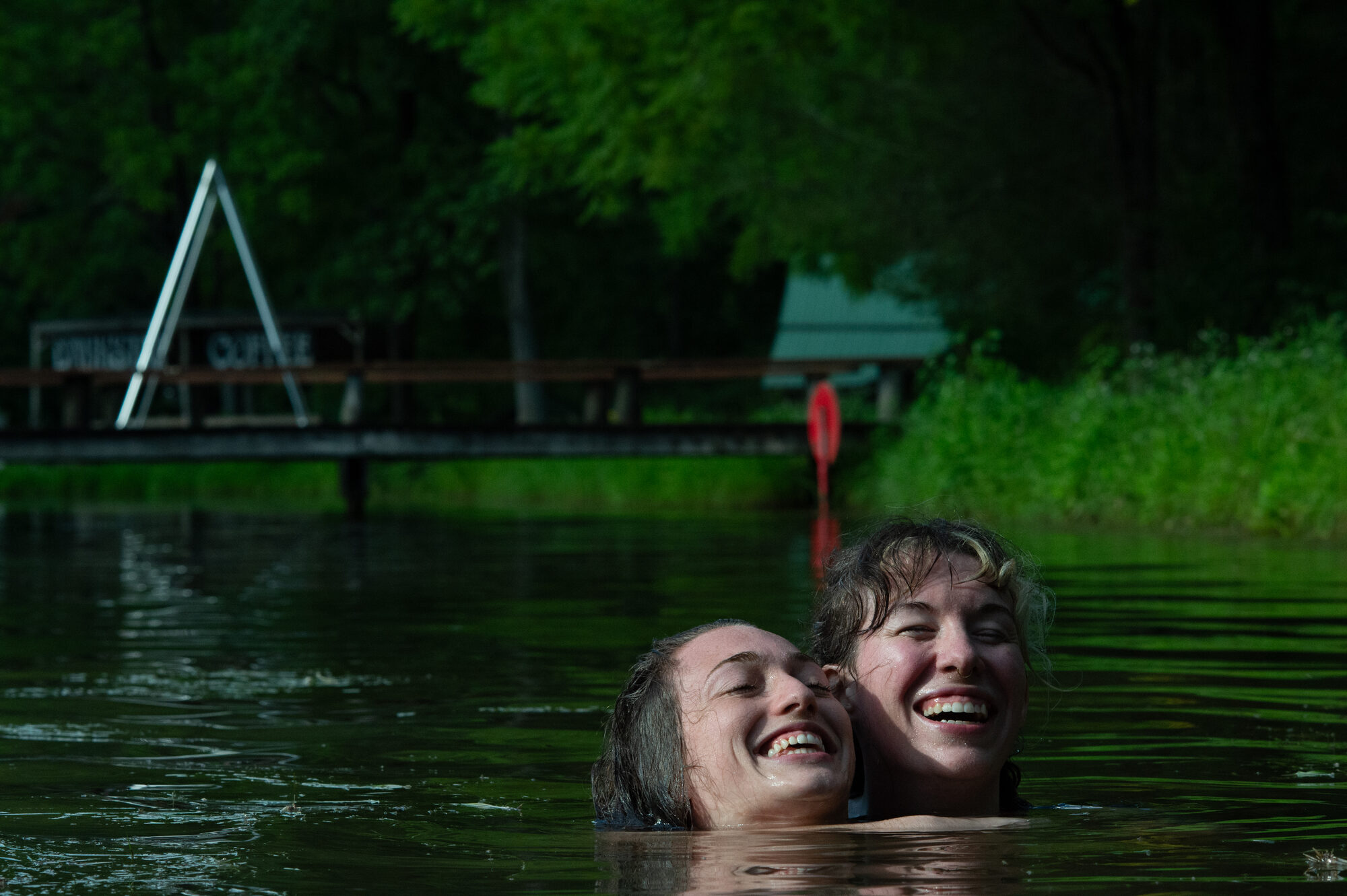Two figures laughing together in a lake with only heads above water, dock and trees in background. Part of a consensual drowning scene with the bottom's legs tied in an agura.