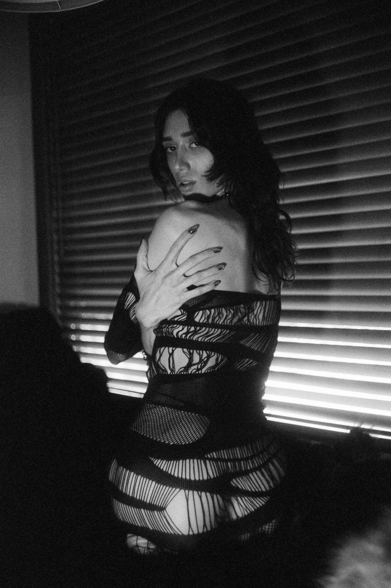 Tate is turning around to face the camera, looking over their back. They are in a distressed, net-like clothing, in front of venetian blinds.