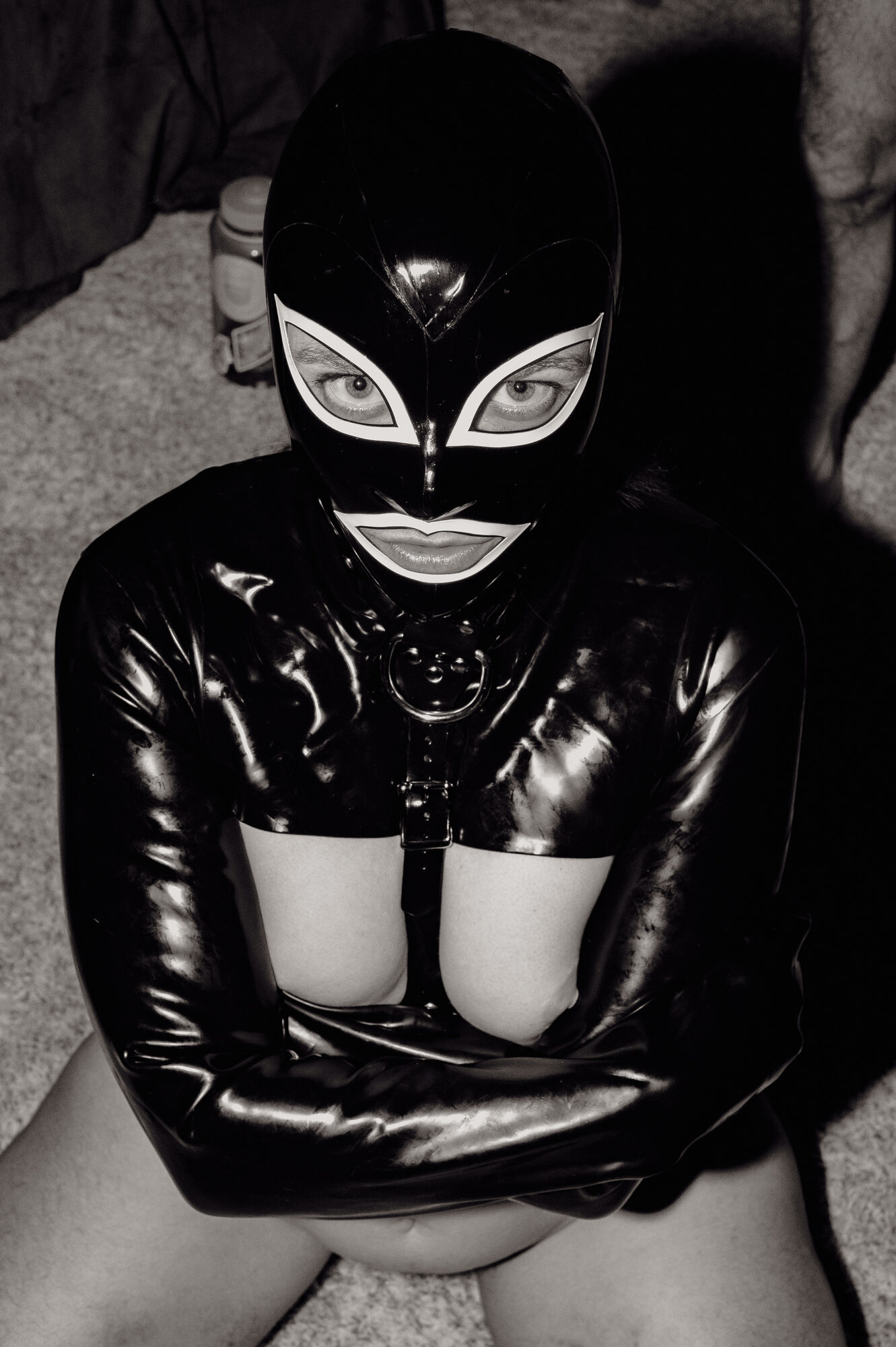Black and white portrait of a figure, E, in a full black latex mask and suit, arms crossed in a heavy latex straight jacket, direct gaze through the eye openings.