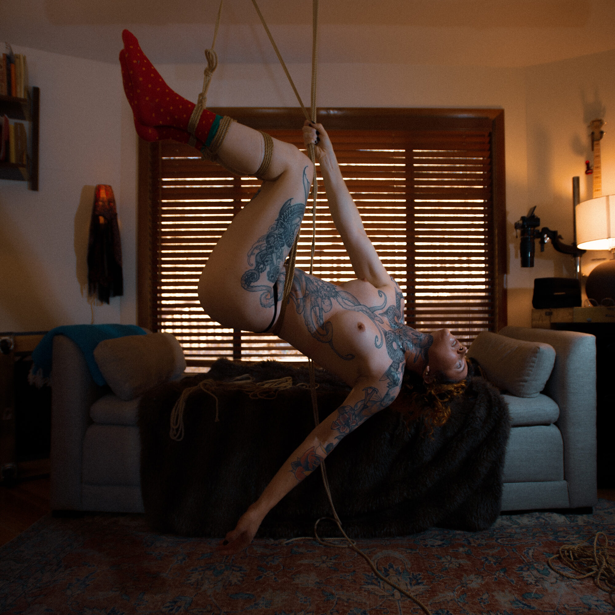 Self portrait. Color rope suspension of a tattooed figure in a warm living room with venetian blinds and lamps.