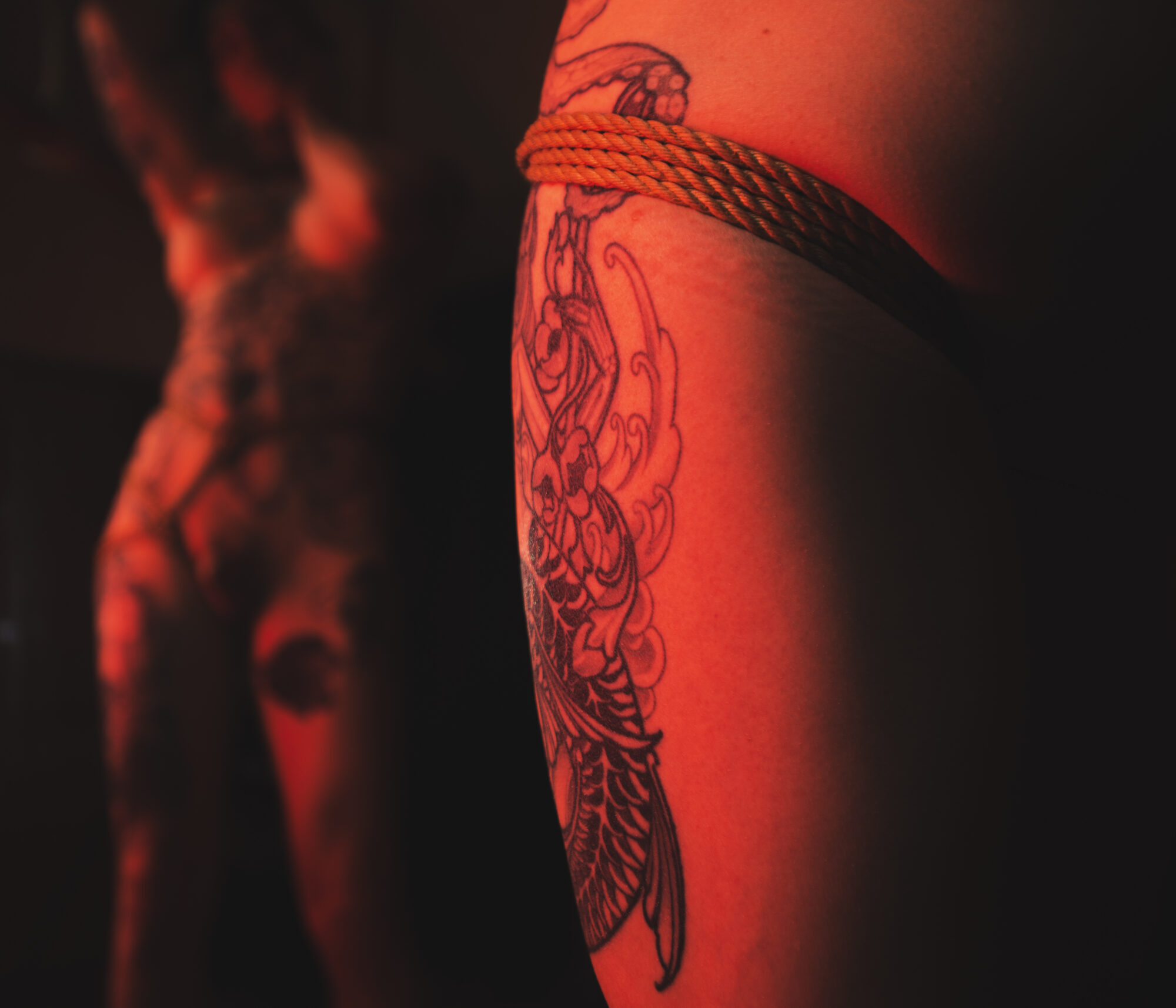 Close-up of tattooed leg with rope binding wrapping under her butt around her thigh in red light, blurred reflection in background.