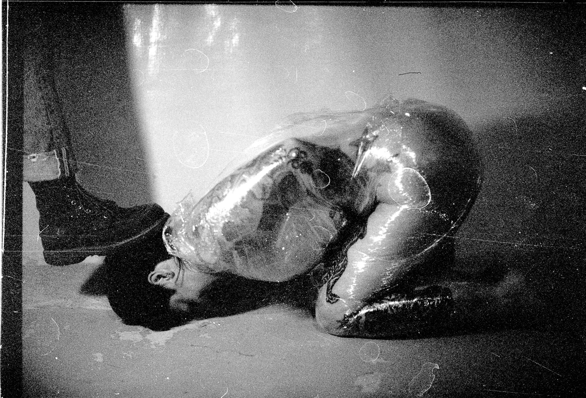 Grainy film picture with lots of imperfections, water spots, black and white. Lauren stands with her booted foot on J's head and they are wrapped in plastic wrap and on their knees, with forehead on the cement ground.