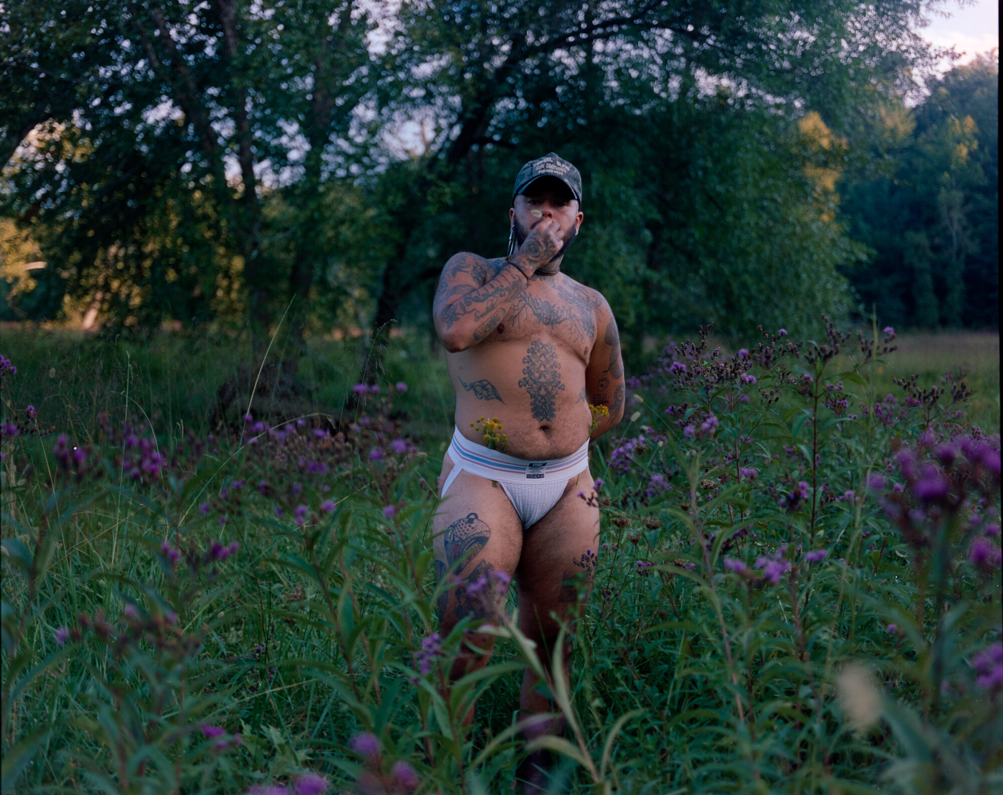 Mo stands waist-deep in ironweed and grass at dusk, hand raised to his mouth, a sprig of goldenrod tucked into the waistband of a white jockstrap. Tattooed torso, camo cap, trees behind. Shot on 120 film.