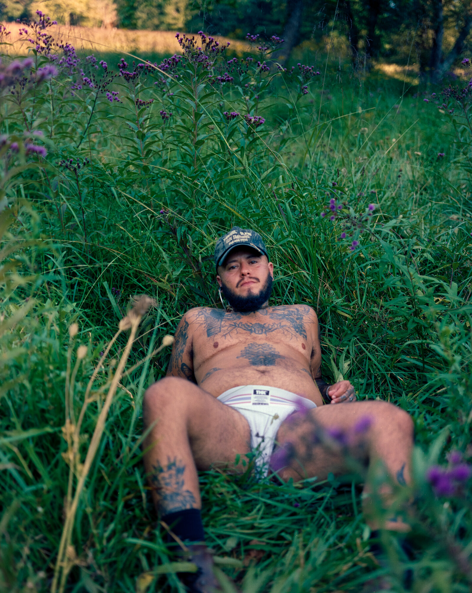 Mo reclines in tall grass looking into the camera, cap on, tattooed chest, white jockstrap and black socks, purple ironweed rising behind him. Shot on 120 film.