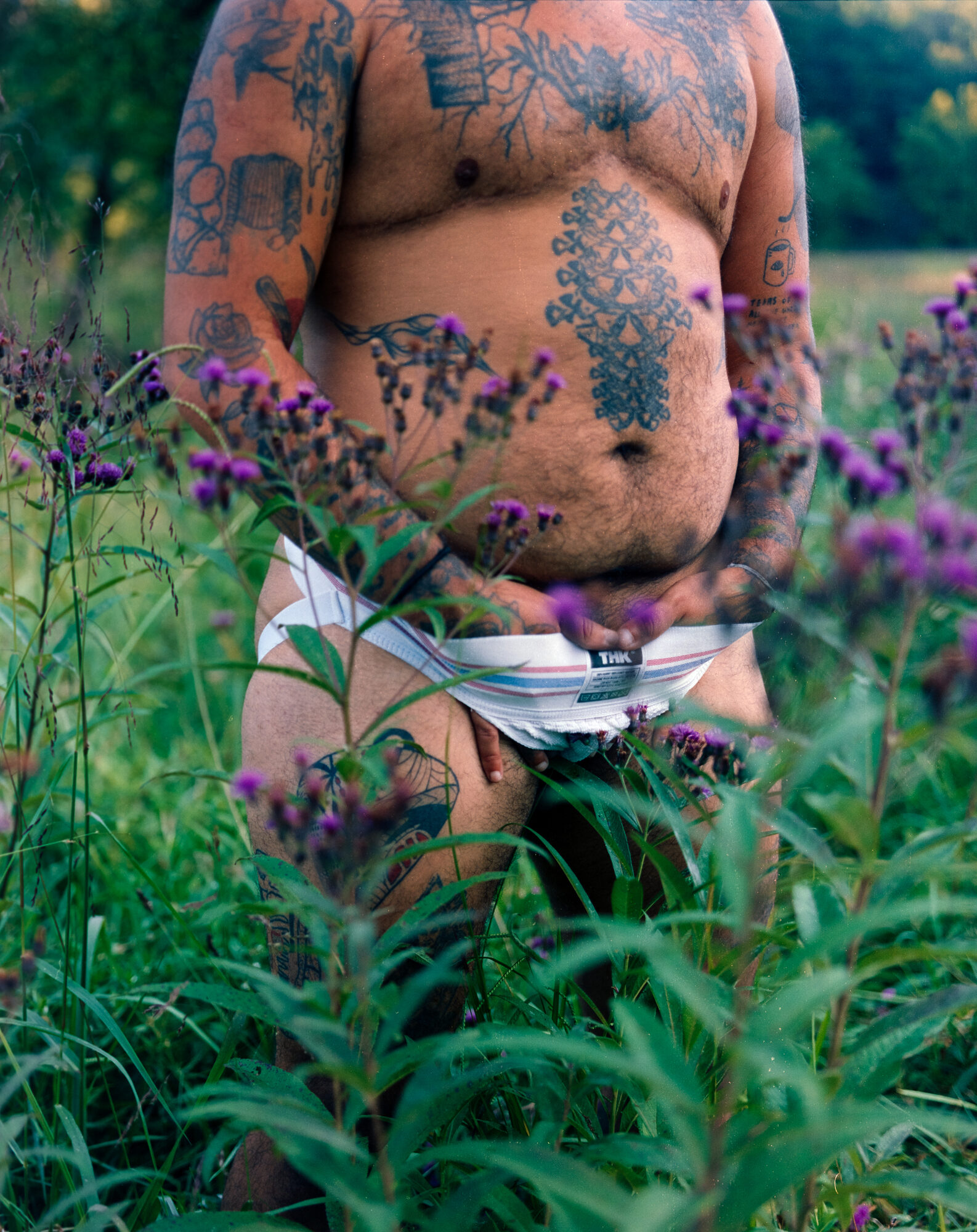 Close view of Mo's tattooed torso and belly, hands resting at the waistband of a white jockstrap, purple ironweed blooms in sharp focus in the foreground. Shot on 120 film.