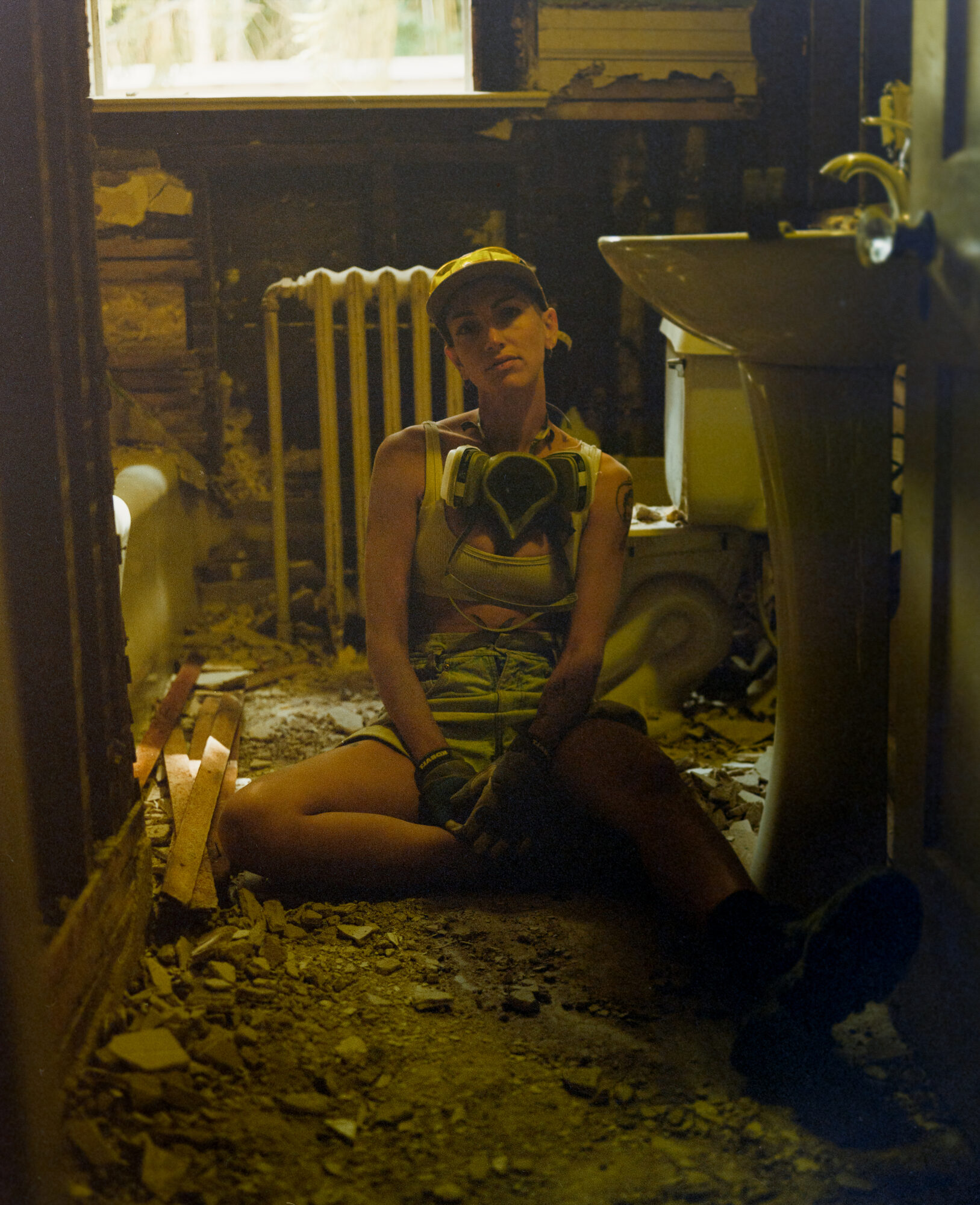 Environmental portrait in a bathroom interior during a remodel. Ryan sits on the floor with a gas mask around her neck, with work boots and gloves sitting in the rubble in warm muted tones. The emotion carries the weight of relationship grief and physical manifestation in the removing of the old and preparing of the new.