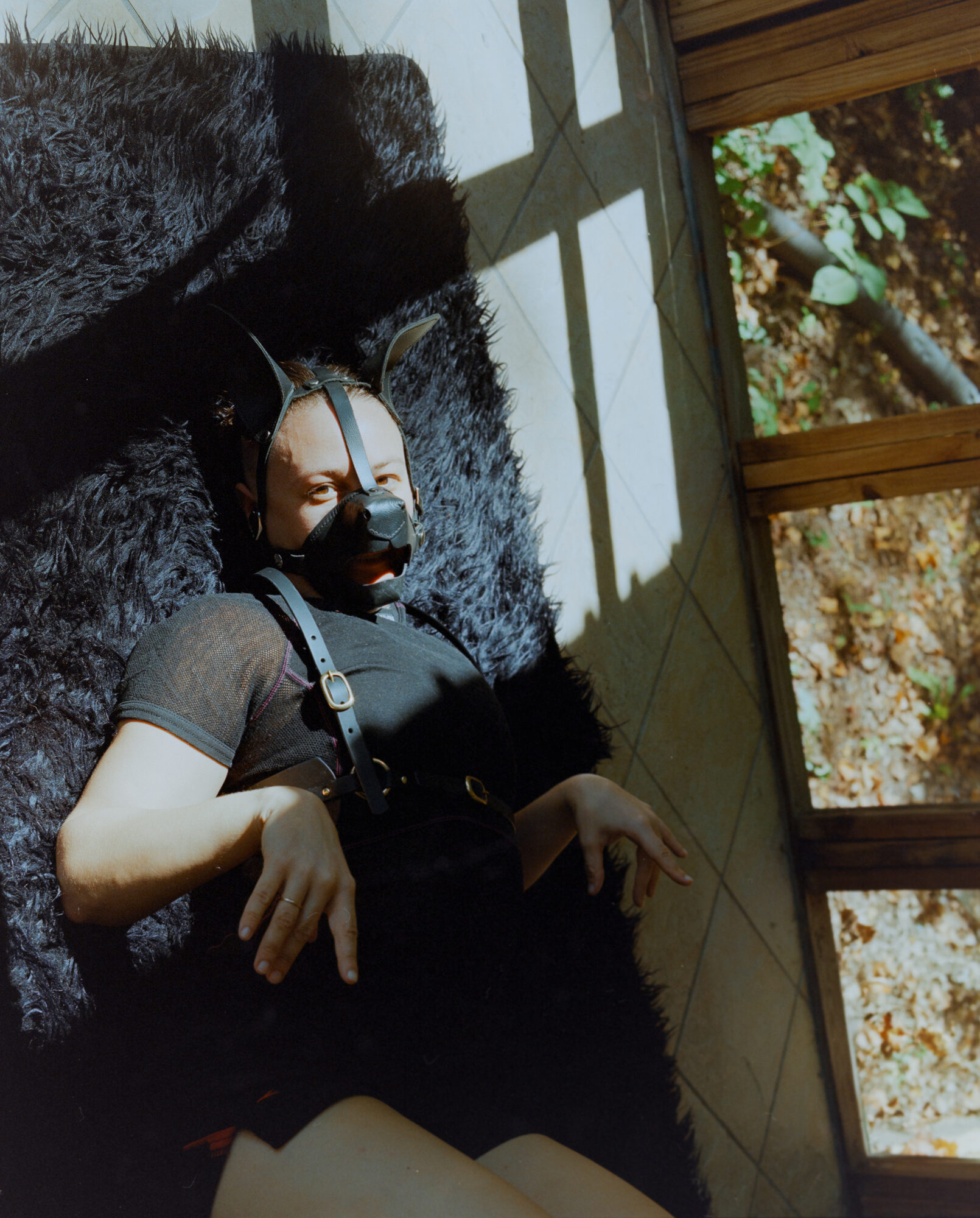 Color portrait of a figure in leather harness and mask reclining against dark fur, dappled sunlight through a window